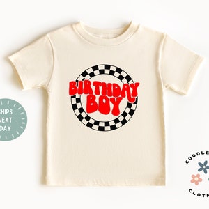 K&ouml;nnte beinhalten: Ein wei&szlig;es T-Shirt mit einer roten "Birthday Boy" Grafik in einer Retro-Schriftart. Die Grafik ist von einem schwarz-wei&szlig; karierten Kreis umgeben.