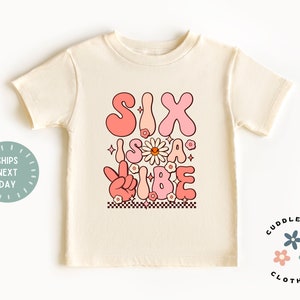 May include: A light beige t-shirt with a pink and white graphic that says "Six is a vibe" with flowers and a peace sign.