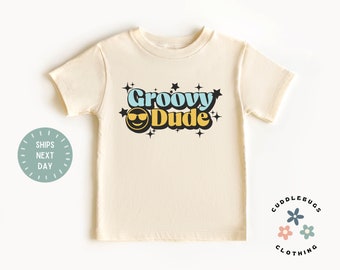 May include: Cream-colored t-shirt featuring the text "Groovy Dude" in blue, yellow, and black, with a smiling face graphic. The shirt has a retro, 70s-inspired design with star accents. The shirt is from Cuddlebugs Clothing.