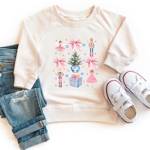 May include: A cream-colored sweatshirt with a Christmas-themed design featuring a Christmas tree, presents, and ballerinas. The design is in shades of pink, blue, and green.