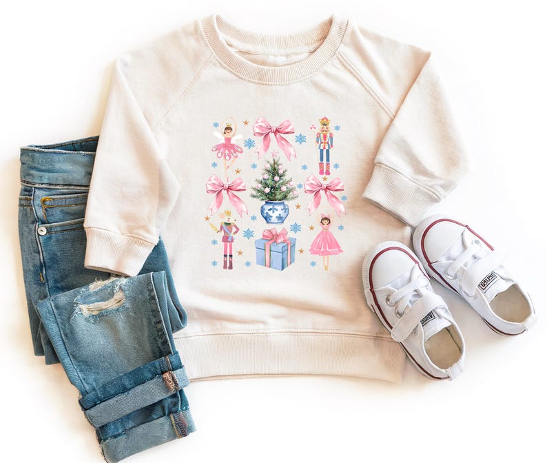 May include: A cream-colored sweatshirt with a Christmas-themed design featuring a Christmas tree, presents, and ballerinas. The design is in shades of pink, blue, and green.