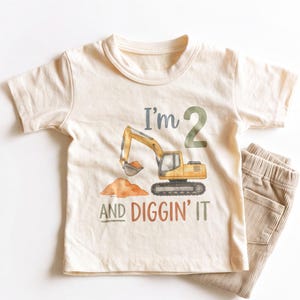 May include: A cream-colored short-sleeve t-shirt featuring a watercolor illustration of a yellow excavator digging into a pile of orange dirt. The text reads I'm 2 AND DIGGIN' IT in dark green and orange lettering, paired with tan corduroy pants.