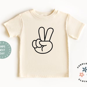 May include: Cream-colored t-shirt featuring a black outline of a hand making the peace sign. The shirt is a short-sleeved crew neck. The words "Cuddlebugs Clothing" are printed on the bottom right.