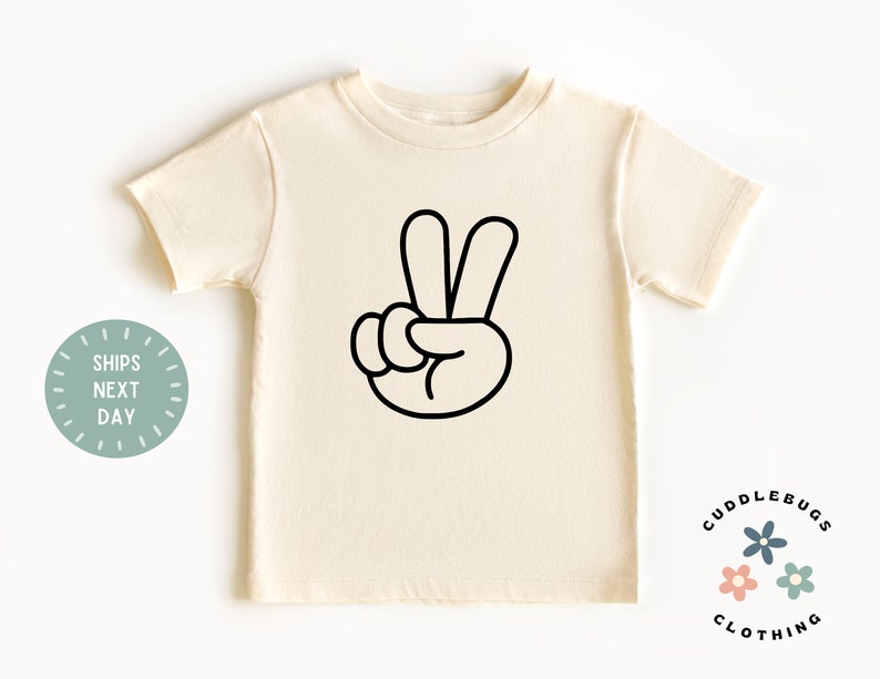 May include: Cream-colored t-shirt featuring a black outline of a hand making the peace sign. The shirt is a short-sleeved crew neck. The words "Cuddlebugs Clothing" are printed on the bottom right.