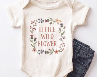 Può includere: Un body a maniche corte color crema con una corona di fiori che circonda la scritta "LITTLE WILD FLOWER" in marrone, abbinato a un paio di jeans in denim grigio antracite scuro con cuciture a vista e bottoni in metallo.
