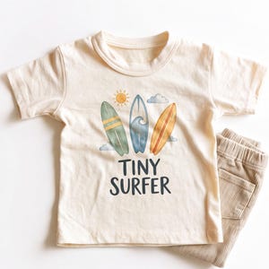 May include: A cream-colored short-sleeve t-shirt featuring a watercolor graphic of three surfboards, a sun, and a cloud. Below the surfboards, the text reads "TINY SURFER" in dark gray letters. The shirt is paired with tan corduroy pants.