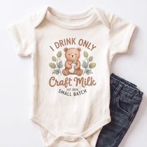 May include: A cream-colored short-sleeve baby bodysuit featuring a graphic of a brown bear holding a baby bottle, surrounded by green hop illustrations. The text reads I DRINK ONLY Craft Milk EST. 2025 SMALL BATCH in brown and black lettering.