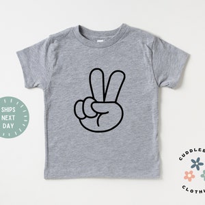May include: A gray short-sleeved t-shirt featuring a black peace sign graphic. The shirt has a crew neck and is made of a soft material. The text "Cuddlebugs Clothing" is printed on the bottom right.