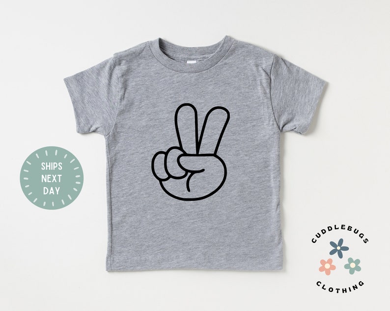May include: A gray short-sleeved t-shirt featuring a black peace sign graphic. The shirt has a crew neck and is made of a soft material. The text "Cuddlebugs Clothing" is printed on the bottom right.