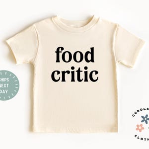 May include: A cream-colored short-sleeved t-shirt with the text "food critic" printed in black.