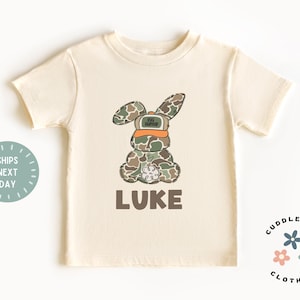 May include: A beige short-sleeved t-shirt with a camouflage bunny wearing a hat that says "Top Hunter" and the name "LUKE" printed below the bunny.