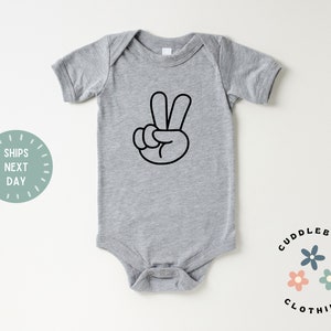 May include: A gray baby onesie with a black peace sign graphic. The onesie has short sleeves and snap closures at the bottom. The text "Cuddlebugs Clothing" and a "Ships Next Day" graphic are also visible.