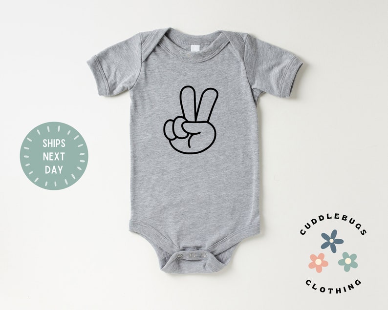 May include: A gray baby onesie with a black peace sign graphic. The onesie has short sleeves and snap closures at the bottom. The text "Cuddlebugs Clothing" and a "Ships Next Day" graphic are also visible.