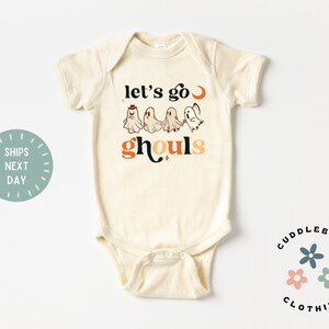 K&ouml;nnte beinhalten: Ein cremefarbener Baby-Strampler mit kurzen &Auml;rmeln zeigt eine Grafik mit Geistern und dem Text "let's go ghouls" in Orange und Schwarz.