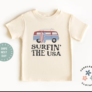May include: A cream colored t-shirt with a vintage van graphic and the text "Surfin' the USA". The van has surfboards on top and an American flag design. The t-shirt has a small logo in the bottom right corner that says "Cuddlebugs Clothing".