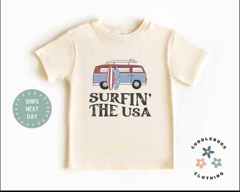 May include: A cream colored t-shirt with a vintage van graphic and the text "Surfin' the USA". The van has surfboards on top and an American flag design. The t-shirt has a small logo in the bottom right corner that says "Cuddlebugs Clothing".