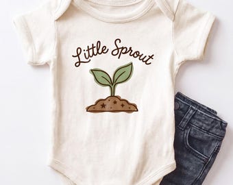 Può includere: Un body a maniche corte color crema con una grafica marrone di una piccola pianta che germoglia dal terreno e la scritta Little Sprout in corsivo marrone sopra la pianta, abbinato a un paio di pantaloni in denim grigio scuro.