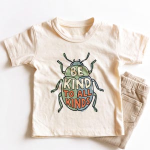 May include: A cream-colored short-sleeve t-shirt featuring a graphic of a beetle with the text BE KIND TO ALL KINDS printed in orange and cream letters on its back, paired with a pair of light brown corduroy pants with an elastic waistband.