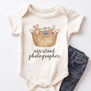 May include: A cream-colored short-sleeve bodysuit featuring a graphic of a woven basket containing a teddy bear, a camera, and a rattle. Below the basket, the text reads assistant photographer in black script. A pair of dark gray jeans is visible.