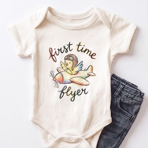 May include: An off-white short-sleeve bodysuit featuring a colorful illustration of a small bird wearing aviator goggles and flying a propeller airplane. The text "first time flyer" is printed in a dark script font above and below the graphic.