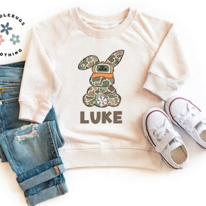 May include: A cream colored sweatshirt with a camouflage bunny wearing an orange hunting hat. The bunny is holding a small white flower. The text "LUKE" is printed below the bunny.