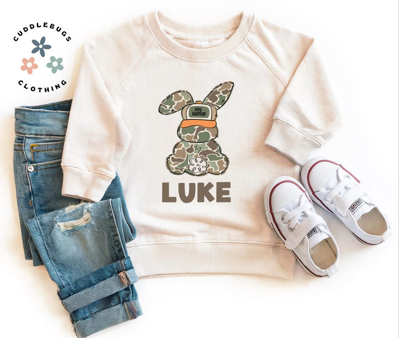 May include: A cream colored sweatshirt with a camouflage bunny wearing an orange hunting hat. The bunny is holding a small white flower. The text "LUKE" is printed below the bunny.