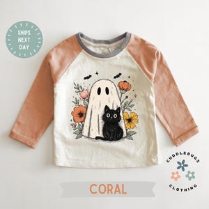 May include: Coral and cream-colored long-sleeved shirt with a Halloween-themed graphic. The graphic features a ghost, a black cat, flowers, and bats. The shirt has coral-colored sleeves and the text "CORAL" at the bottom.