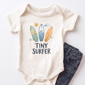 May include: An off-white short-sleeve bodysuit featuring a graphic of three surfboards in green, blue, and orange, a yellow sun, and gray clouds. Below the surfboards, the text reads "TINY SURFER" in dark gray letters. Dark gray denim pants are included.