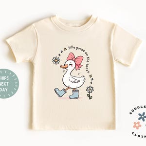 May include: A beige short-sleeved t-shirt with a cartoon illustration of a goose wearing pink boots and a pink bow. The text "Silly goose on the loose" is written in a circle around the goose.