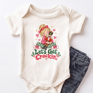 May include: An off-white short-sleeve bodysuit featuring a graphic of a blonde person with a pink bow holding a nutcracker. The design includes green plaid pants, pink hearts, and the text Let's Get Crackin' in green and red with holly accents.