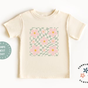 May include: Cream-colored t-shirt featuring a square design with pink flowers and a green and white checkered background. The shirt has short sleeves and a crew neck. The text "SHIPS NEXT DAY" is on the left side of the shirt.