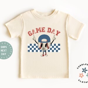 May include: Cream-colored t-shirt featuring a baseball character wearing a blue cap, with the words "GAME DAY" in red. The baseball character is pointing upwards. A blue and white checkered pattern is below the character.