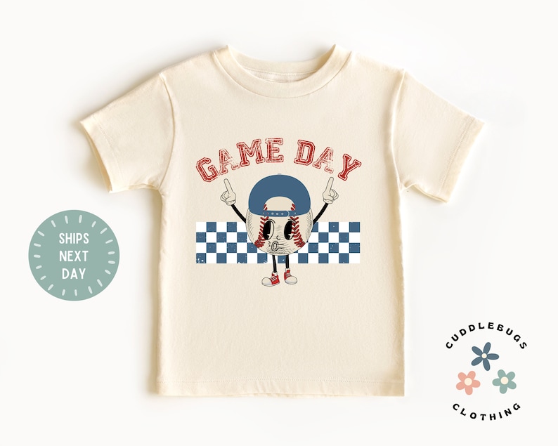 May include: Cream-colored t-shirt featuring a baseball character wearing a blue cap, with the words "GAME DAY" in red. The baseball character is pointing upwards. A blue and white checkered pattern is below the character.