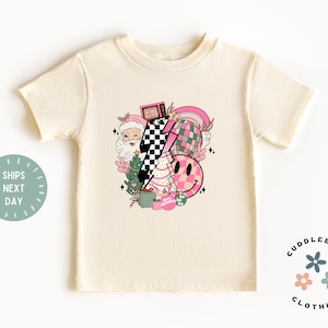 K&ouml;nnte beinhalten: Ein cremefarbenes T-Shirt mit kurzen &Auml;rmeln und einem pinkfarbenen und gr&uuml;nen Weihnachtsmotiv. Das Motiv zeigt den Weihnachtsmann, einen Weihnachtsbaum, Geschenke, ein Schachbrettmuster, ein Smiley und einen Regenbogen. Der Text "Home Alone" ist auf einem Fernseher im Motiv zu sehen.
