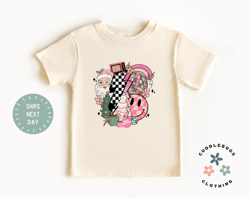 K&ouml;nnte beinhalten: Ein cremefarbenes T-Shirt mit kurzen &Auml;rmeln und einem pinkfarbenen und gr&uuml;nen Weihnachtsmotiv. Das Motiv zeigt den Weihnachtsmann, einen Weihnachtsbaum, Geschenke, ein Schachbrettmuster, ein Smiley und einen Regenbogen. Der Text "Home Alone" ist auf einem Fernseher im Motiv zu sehen.