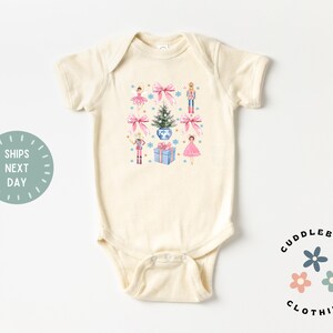 May include: A cream-colored baby bodysuit with a Christmas-themed design featuring a Christmas tree, presents, a nutcracker, and ballerinas. The design is in shades of pink, blue, and green.