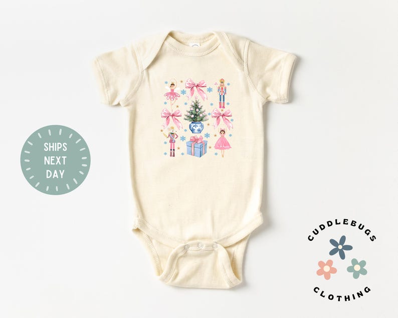 May include: A cream-colored baby bodysuit with a Christmas-themed design featuring a Christmas tree, presents, a nutcracker, and ballerinas. The design is in shades of pink, blue, and green.