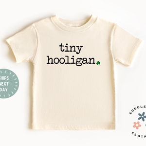 May include: A cream colored short-sleeved t-shirt with the text "tiny hooligan" in black lettering and a green shamrock.
