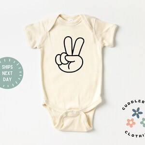 May include: Cream-colored baby onesie with a black peace sign graphic. The onesie has short sleeves and a round neckline. The text "Cuddlebugs Clothing" is printed in black, along with three small flower graphics.