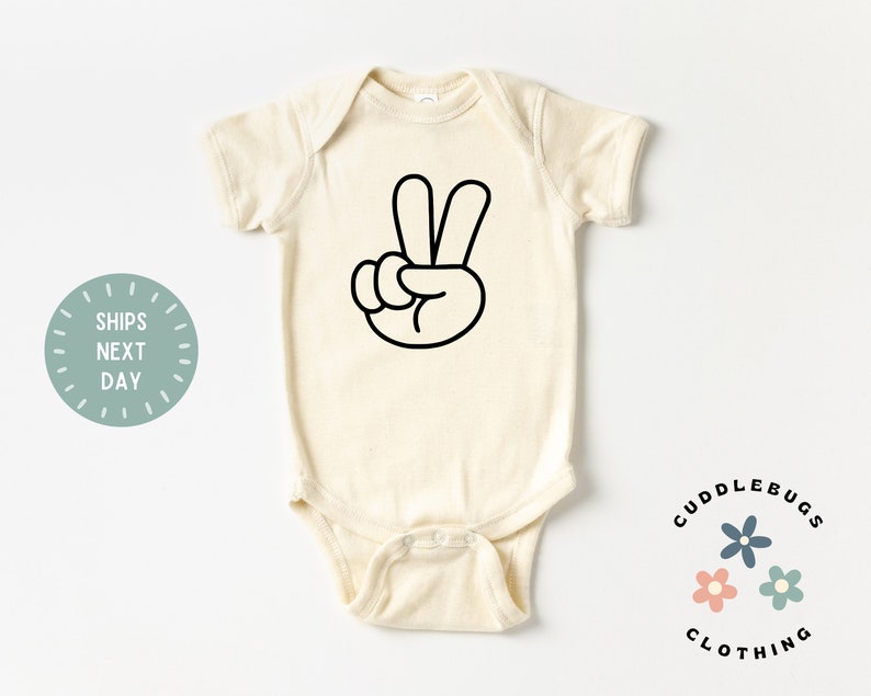 May include: Cream-colored baby onesie with a black peace sign graphic. The onesie has short sleeves and a round neckline. The text "Cuddlebugs Clothing" is printed in black, along with three small flower graphics.