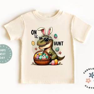 May include: A light beige t-shirt with a graphic of a dinosaur wearing sunglasses and bunny ears, holding a basket of Easter eggs. The text "On the Hunt" is printed on the shirt.