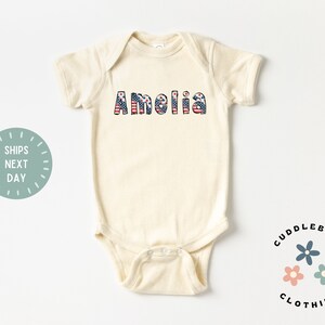 May include: A cream-colored baby onesie with the name "Amelia" printed in red, white, and blue, resembling the American flag.