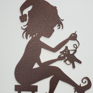 Könnte beinhalten: Eine kupferfarbene Metallsilhouette eines Elfen, der auf einem Hocker sitzt und näht. Der Elf trägt einen Hut und hat langes Haar. Der Elf hält in der einen Hand eine Nadel und Faden und in der anderen ein Stück Stoff.