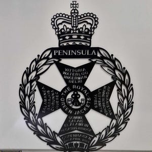 Peut inclure: Emblème en métal noir et blanc avec une couronne en haut et une couronne de laurier autour. L'emblème représente une croix avec les mots "The Royal Green Jackets" et les noms de batailles et de lieux, notamment "Peninsula", "Vittoria", "Waterloo", "Inkerman", "Delhi", "Salamanca", "Badajos", "Corunna", "Quebec", "Somme", "Calais", "El Alamein", "Pegasus Bridge", "Copenhague 2 avril 1801", "Ypres", "Ladysmith", "Nonne Bosschen" et "Afghanistan".