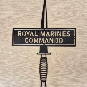 May include: Black metal wall art featuring a stylized dagger with a serrated blade and a textured handle. The dagger is positioned above a rectangular sign that reads "ROYAL MARINES COMMANDO".