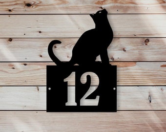 May include: Black metal house number sign with a silhouette of a cat looking up. The number 12 is displayed in white on the sign.