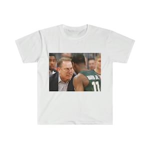 May include: A white t-shirt featuring a photograph of a basketball coach speaking to a player. The player is wearing a green and white basketball jersey with the number 11 on it. The text on the jersey reads "NORTH DE".