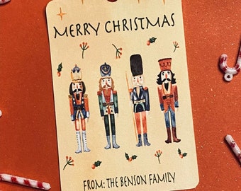Peut inclure: Une étiquette cadeau de Noël beige avec le texte "Joyeux Noël" et quatre illustrations de casse-noisettes. L'étiquette comprend également le texte "De la part de la famille Benson".
