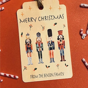May include: A beige Christmas gift tag with the text "Merry Christmas" and four nutcracker illustrations. The tag also includes the text "From: The Benson Family".