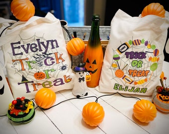 Könnte beinhalten: Zwei weiße Stoff-Trick-or-Treat-Taschen mit den Namen Evelyn und Elijah bedruckt. Die Taschen sind mit Halloween-Motiven verziert. Auf einer weißen Holzoberfläche liegen mehrere orangefarbene Kürbisse und eine Geisterfigur.